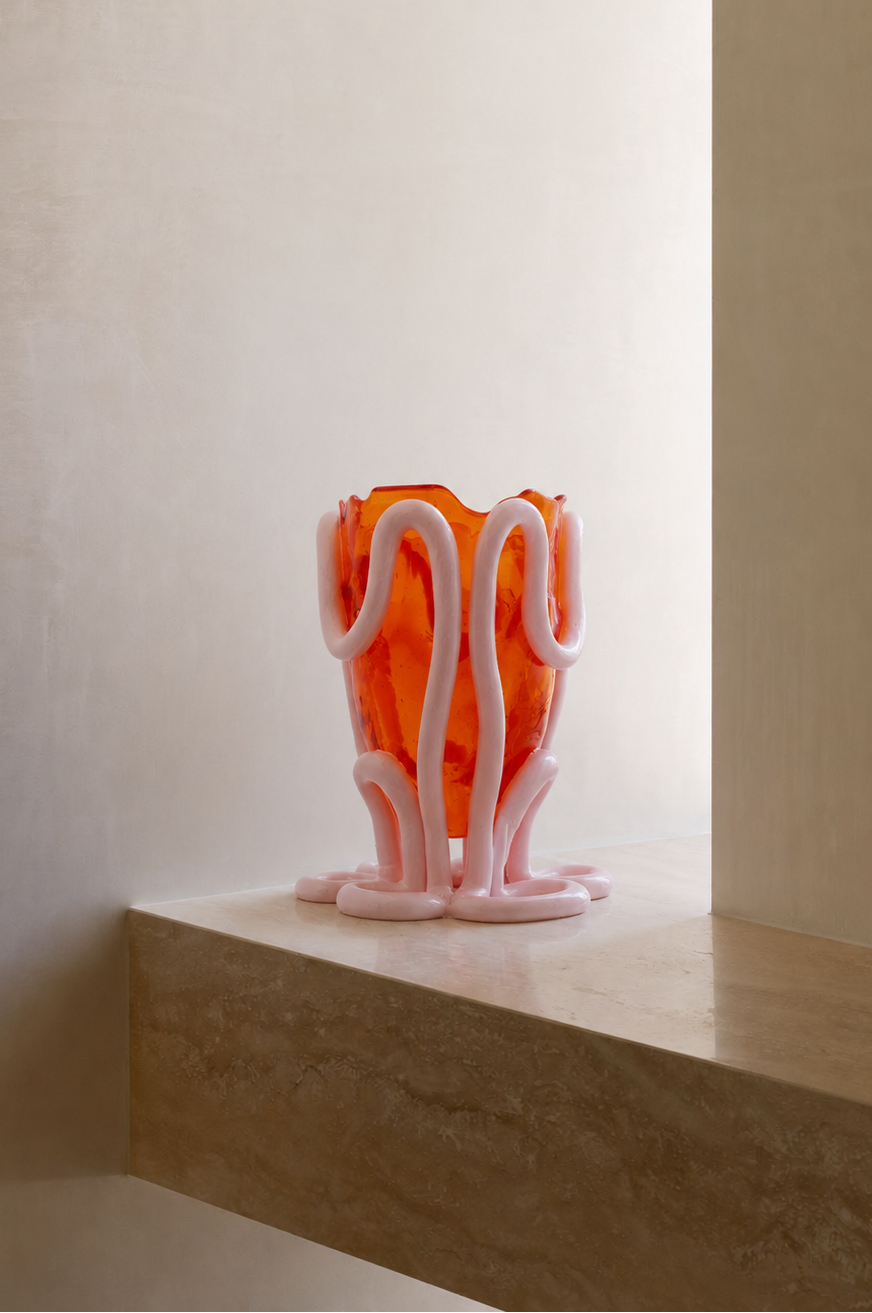 A sculptural glass vase with a wavy, organic pink form and translucent orange interior, displayed on a stone ledge against a warm beige wall.